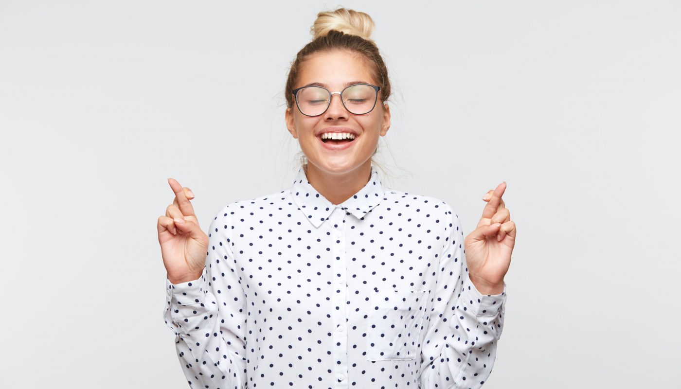Closeup of cheerful young woman with bun and closed eyes wears polka dot shirt and glasses feels inspired, keeps fingers crossed and making a wish isolated over white background